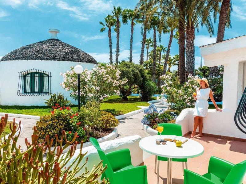 Outdoor seating area with green chairs and round table near white round building and palm trees.