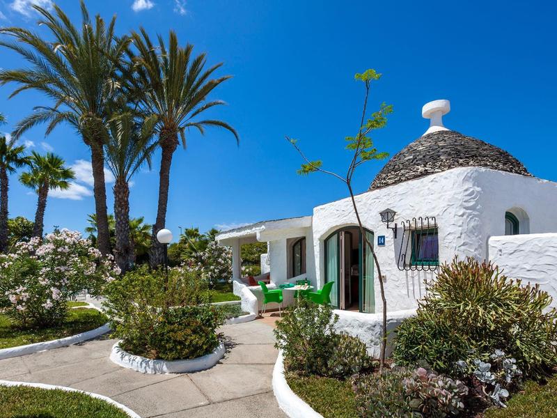White stone villa with round roof surrounded by palm trees and lush garden.