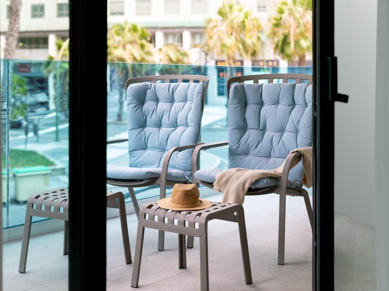 Balcón con dos sillones azules, reposapiés, un sombrero y vista a palmeras y área de piscina.