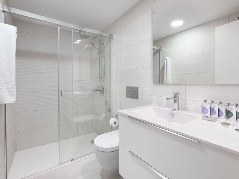 Baño moderno y luminoso con ducha de vidrio, lavabo blanco y espejo grande.