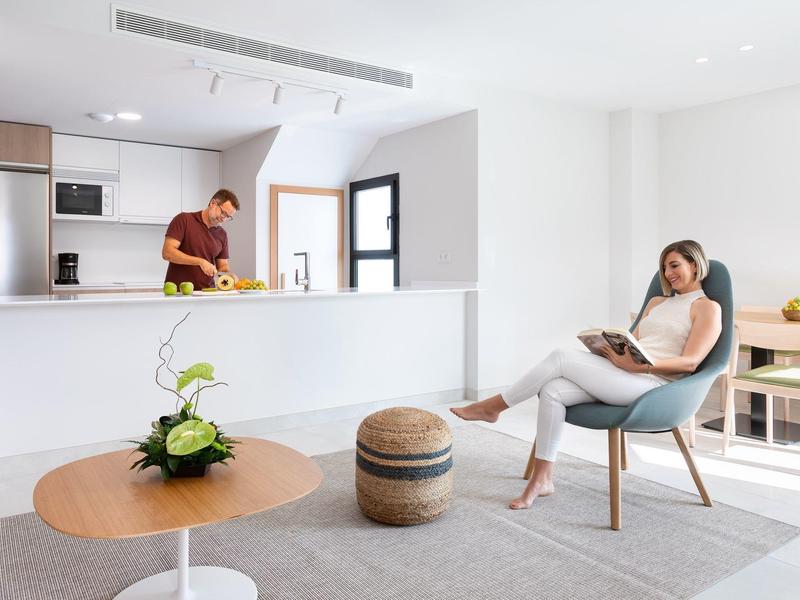 Apartamento moderno y luminoso con cocina abierta, hombre en la cocina y mujer relajada en una silla.