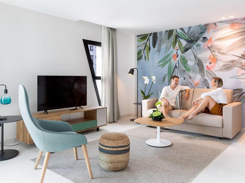 Habitación de hotel moderna y luminosa con una pareja en el sofá frente a una pared con diseño colorido.