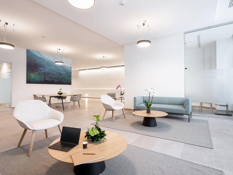 Área de salón de hotel moderna y luminosa con sillas blancas, sofá gris y varias mesas.