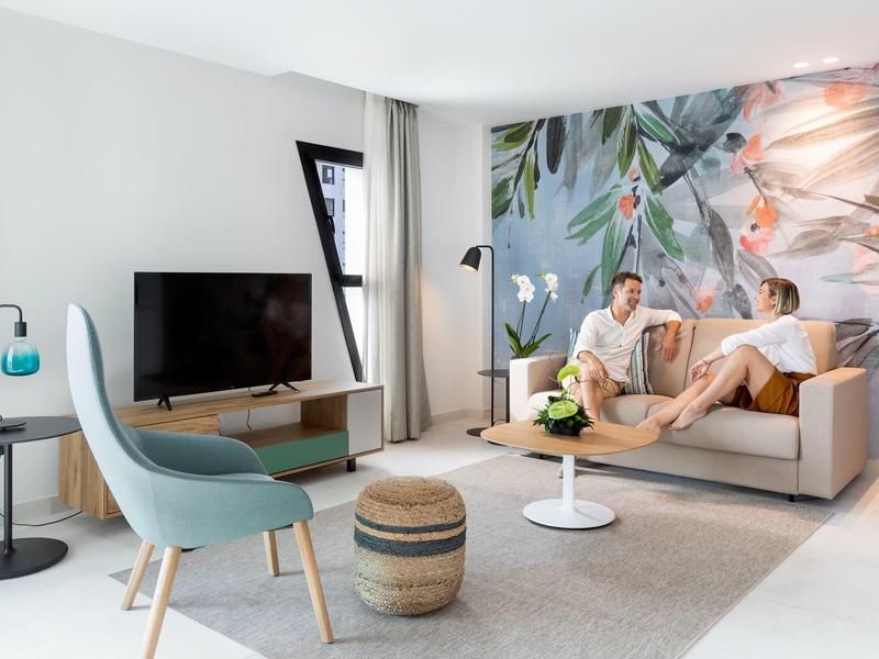 Sala moderna amueblada con sofá, decoración floral en la pared y televisión para relajarse en la habitación del hotel.