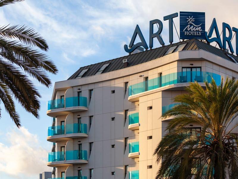 Edificio de hotel moderno con balcones azules y palmeras en primer plano a plena luz del día.