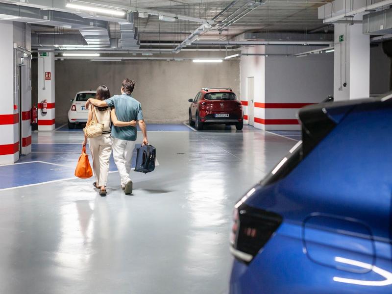 Dos personas caminan en un estacionamiento subterráneo hacia autos estacionados, una lleva bolsas de compras.