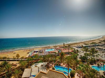 Strandhotel mit mehreren Pools, Palmen und weiter Sicht aufs Meer bei klarem Himmel.
