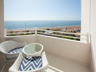 Balkon s bílými židlemi, skleněným stolkem a výhledem na moře za slunečného dne