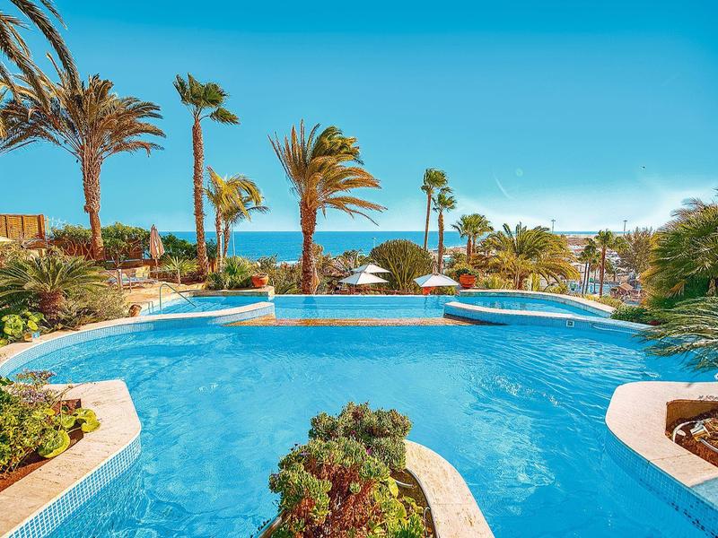 Großer Hotelpool mit Palmen und Meerblick unter klarem Himmel.