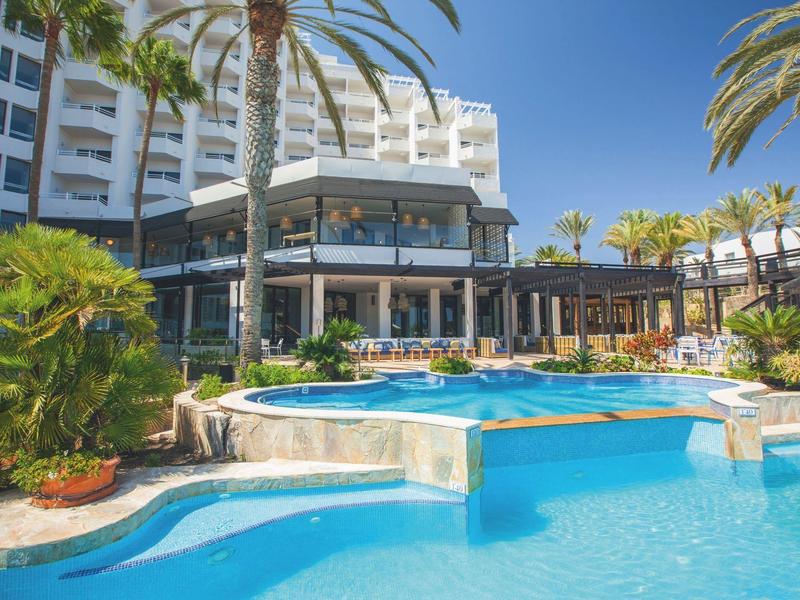 Hotel mit rundem Pool und Palmen bei klarem Himmel, umgeben von blühenden Pflanzen.