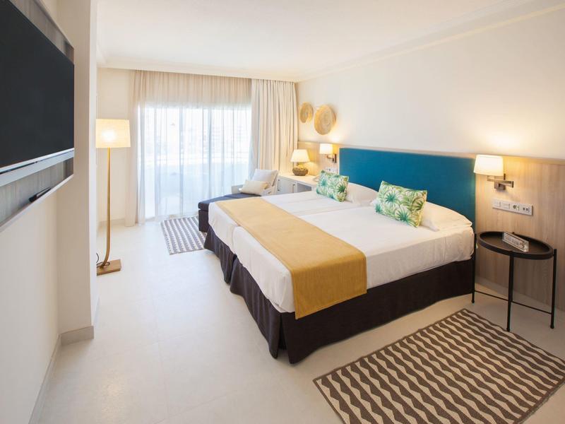 Hell eingerichtetes Hotelzimmer mit zwei Betten, blauem Kopfteil und Balkonlicht.