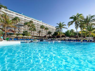 Large hotel pool with palm trees and lounge chairs under a clear blue sky.