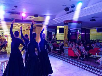 Four dancers in black dresses perform a choreography in front of a seated audience.