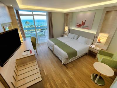 Moderne hotelkamer met groot bed, tv, balkon, groene fauteuil en houten vloer.