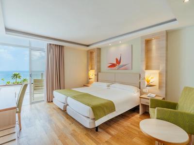 Nowoczesny pokój hotelowy z podwójnym łóżkiem, drewnianą podłogą i zielonym fotelem przy balkonie z widokiem na morze.