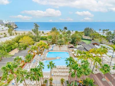 Widok na basen hotelowy z palmami i oceanem w tle w słoneczny dzień.