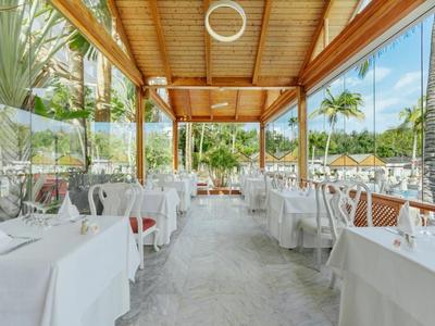 Otwarta zadaszona strefa restauracyjna hotelu z białymi obrusami i widokiem na basen i palmy.