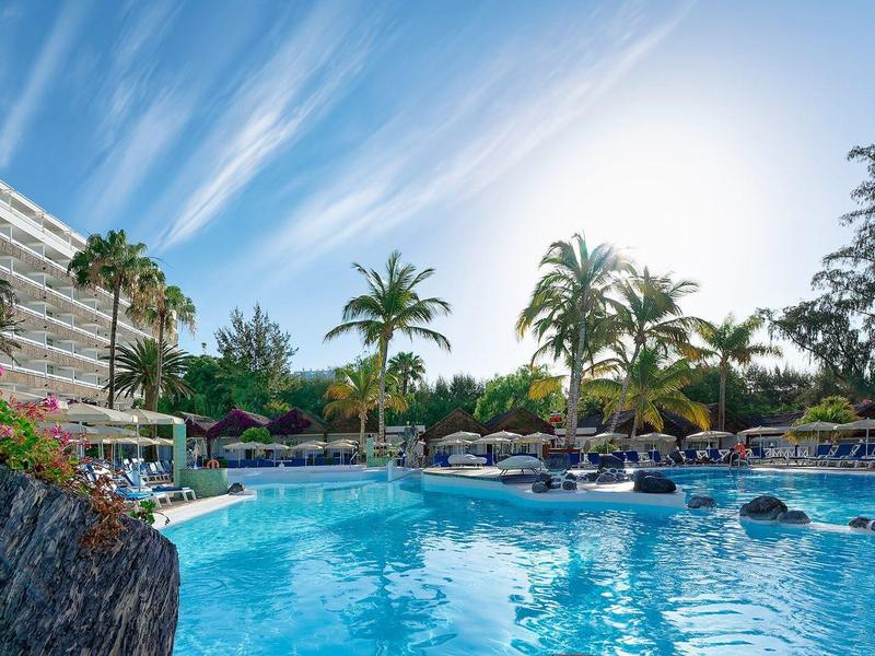 Resortzwembad omgeven door palmbomen, ligstoelen en een modern hotelgebouw onder een heldere hemel.