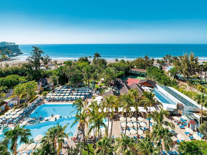 Hotelresort met zwembaden, palmbomen en uitzicht op zee en groene heuvels.