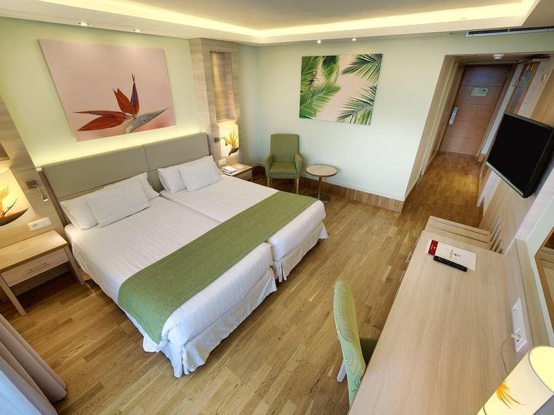 Moderne hotelkamer met twee eenpersoonsbedden, groene accenten, houten vloer, bureau en fauteuil.