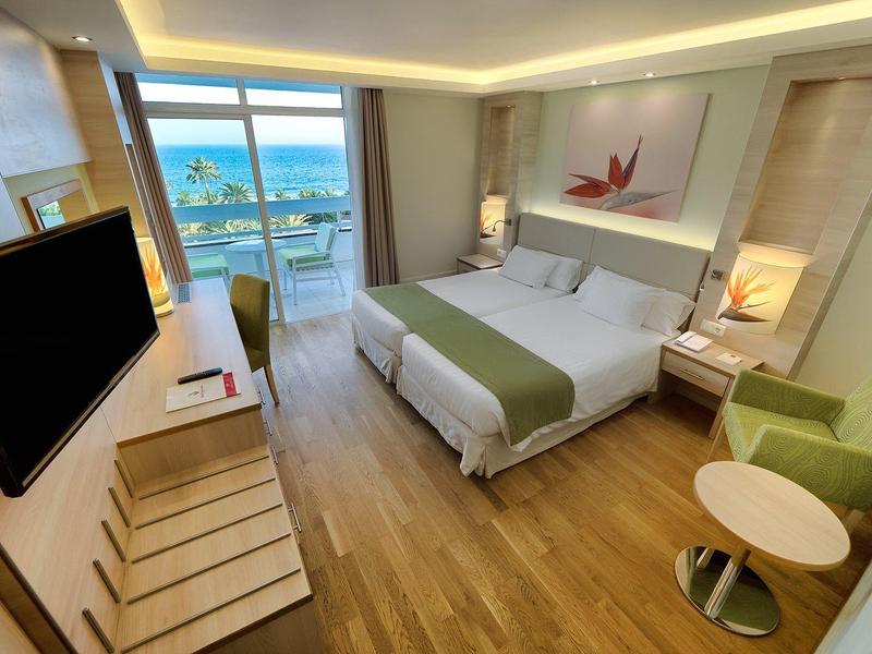 Moderne hotelkamer met groot bed, tv, balkon, groene fauteuil en houten vloer.