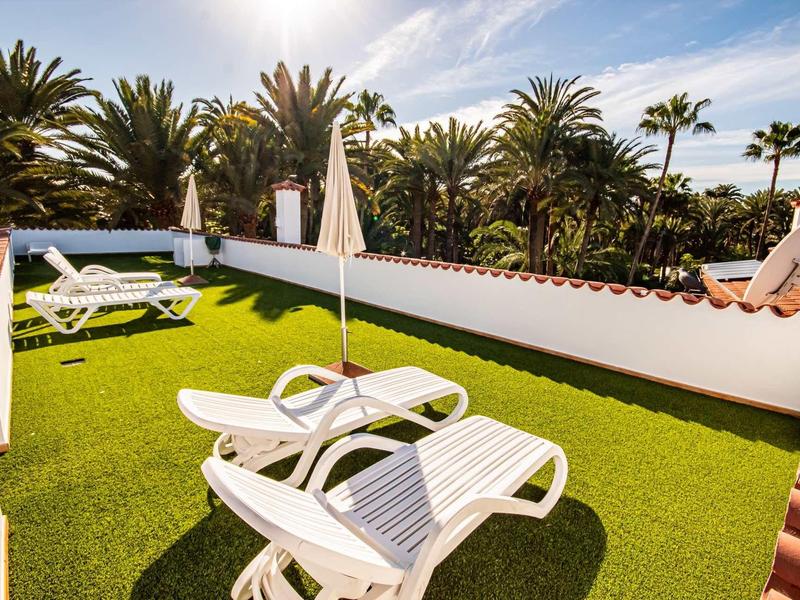 Taras słoneczny z białymi leżakami, zieloną sztuczną trawą i palmami w tle.