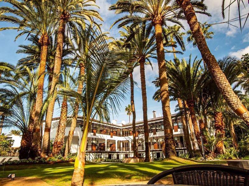 Hotel z wieloma wysokimi palmami i brukowaną ścieżką na pierwszym planie w słoneczny dzień.