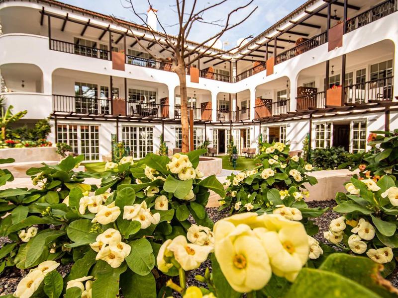 Dziedziniec hotelu z kwitnącymi żółtymi kwiatami i białymi balkonami na dwóch piętrach.