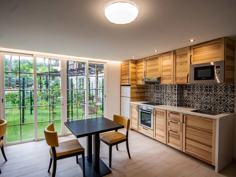 Nowoczesna kuchnia z drewnianymi meblami i jadalnią, duże okna z widokiem na zielony ogród.