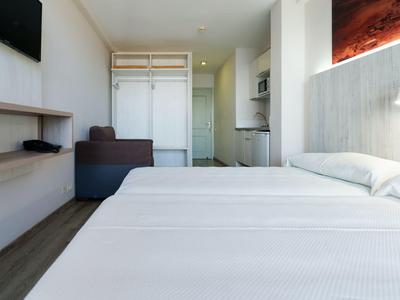 Nowoczesny pokój hotelowy z łóżkiem podwójnym, sofą, otwartą garderobą i małym aneksem kuchennym w neutralnych kolorach.