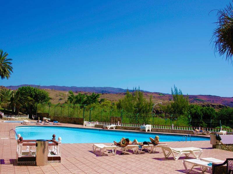 Basen na świeżym powietrzu z leżakami i palmami na tle gór.