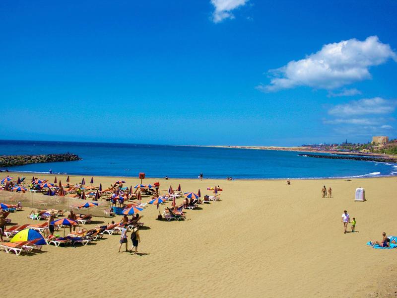 Szeroka piaszczysta plaża z ludźmi wypoczywającymi w słońcu pod jasnym błękitnym niebem.