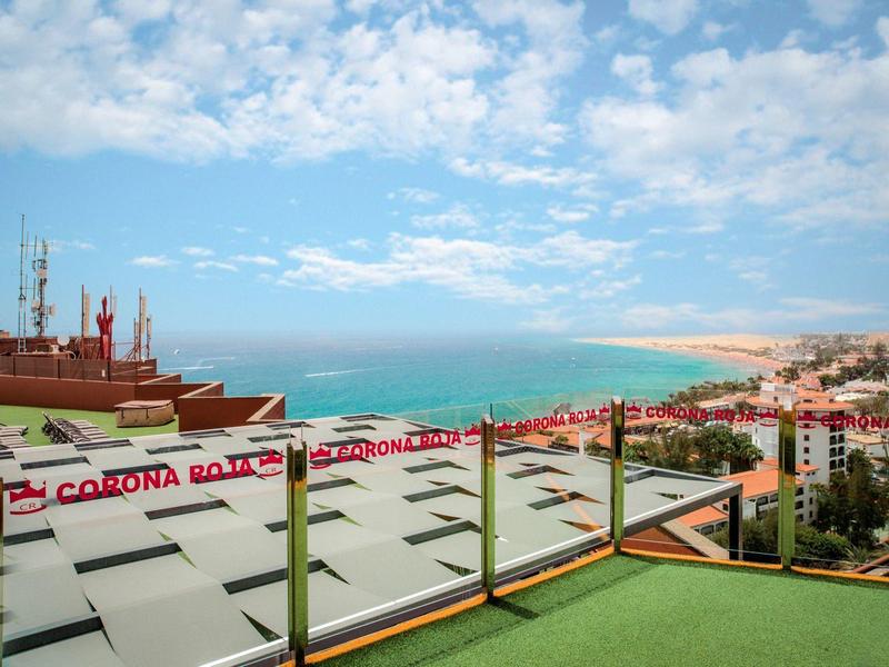 Widok z tarasu na dachu z przeszkloną balustradą na morze i piaszczystą plażę pod zachmurzonym niebem.