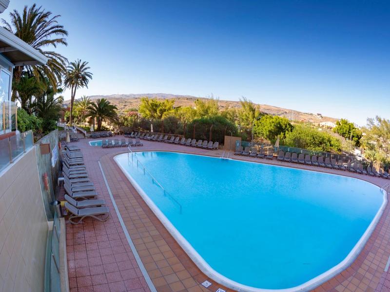 Basen na świeżym powietrzu z leżakami, otoczony palmami pod błękitnym niebem, otoczenie hotelowe.