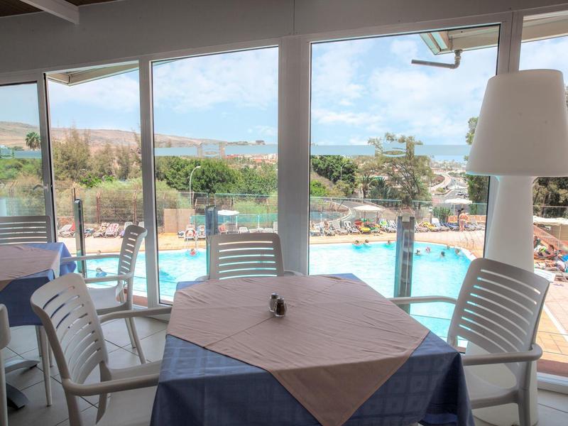 Widok przez duże okna na basen z stołami i białymi krzesłami w restauracji.