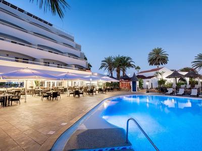 Hotelterrasse mit beleuchtetem Pool, Sitzgruppen und Palmen bei klarem Himmel am Abend.