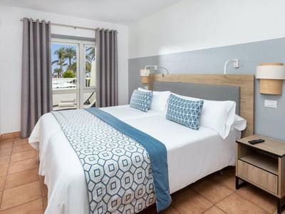 Chambre d'hôtel lumineuse avec lit double, table de chevet et balcon avec vue sur des palmiers.