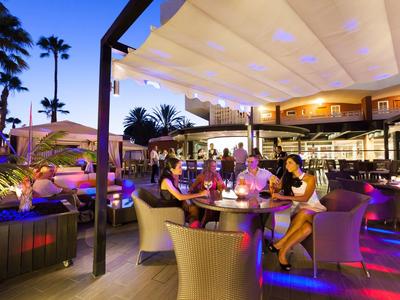 Scène en soirée avec des clients assis sous une terrasse couverte d'un hôtel tropical.