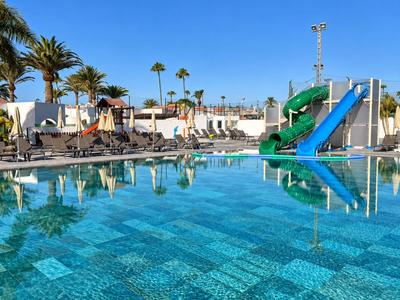 Klare blaue Hotelpoollandschaft mit Wasserrutschen, Sonnenliegen und Palmen bei strahlendem Himmel.