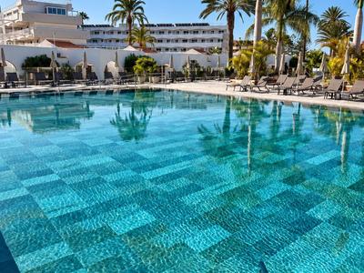 Klares Schwimmbad mit Fliesen umgeben von Liegestühlen und Palmen vor einem Hotel.