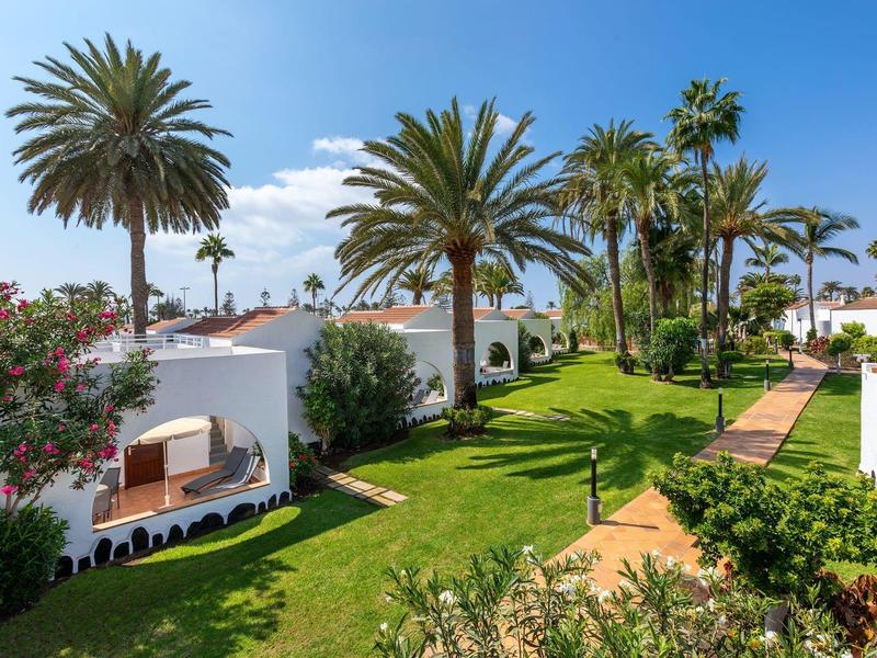 Bungalows blancs avec des chemins en herbe entourés de palmiers et d'une végétation luxuriante sous un ciel bleu.