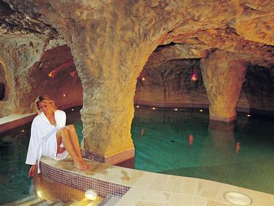 Unterirdisches Bad mit Felsenwänden, großem Wasserbecken und Frau im Bademantel auf Treppenstufe.