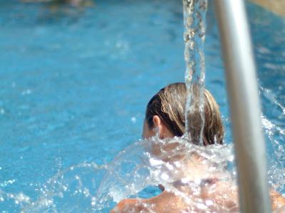 Mann mit nassem Rücken unter Wasserdusche am Pool, klare Wasserstrahlen, grün-helle Umgebung.