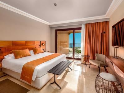 Helles Hotelzimmer mit großem Bett, orangen Akzenten und Balkon mit Meerblick.