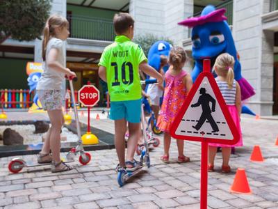 Kinder mit Rollern stehen auf einem Hof, ein Hundemaskottchen begrüßt sie, Verkehrszeichen sind sichtbar.