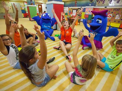 Kinder sitzen im Kreis auf gestreiftem Boden und spielen mit zwei blauen Maskottchen.