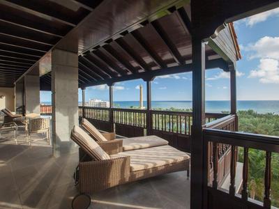 Balkon mit Liegestuhl und Stühlen, Holzdach, Blick auf Meer und grünen Wald bei blauem Himmel.