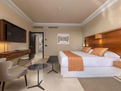 Modernes Hotelzimmer mit Doppelbett, Holzmöbeln, beige Wänden und Sitzbereich mit Sesseln und Tischen.