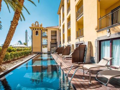 Moderner Hotelpool mit Liegestühlen, Palmen und gelbem Gebäude bei klarem Himmel.
