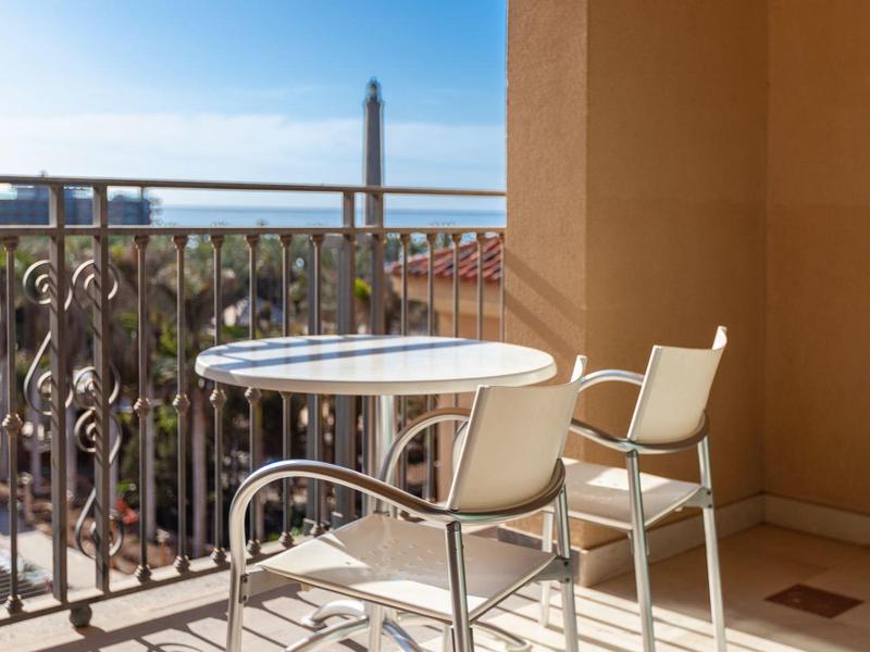 Balkon se stolem a dvěma židlemi, výhled na palmy a věž za jasného nebe.